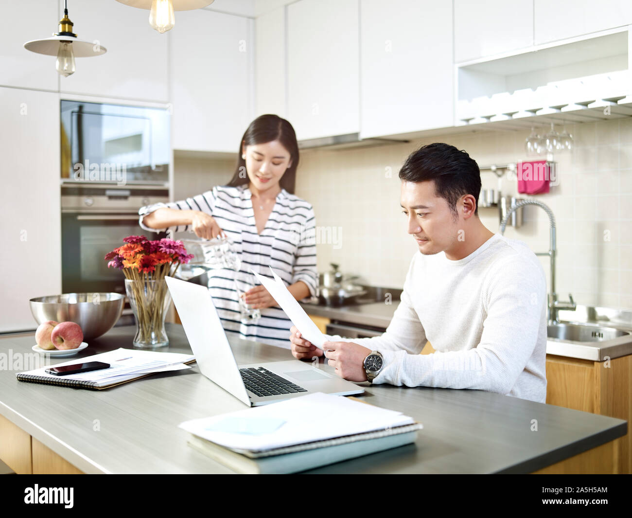 young asian man working at home using laptop computer while wife pouring a glass of water Stock Photo