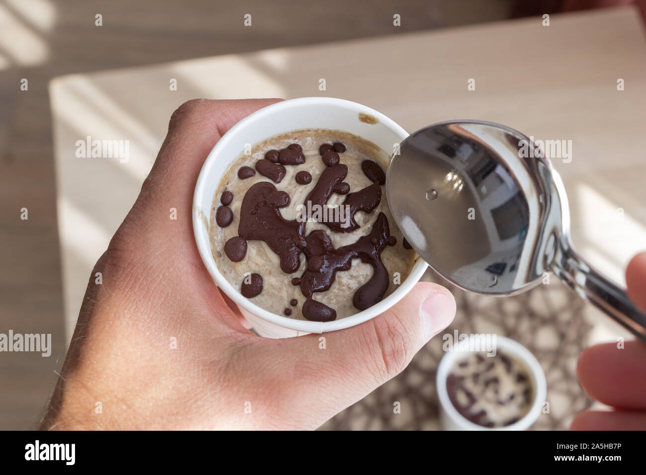 A men eats healthy raw ice cream with spoon. Top view from myself. delicious fruit vegan dessert in a white paper cup in a male hand. A small spoon in Stock Photo