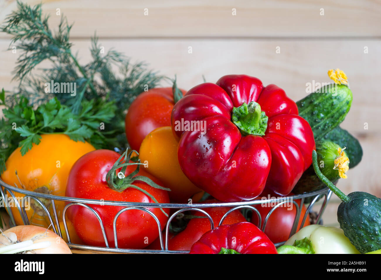 Close up vegetables harvest in wicker basket on wooden background. Red and yellow pepper, tomatoes, onion and green bow, parsley, dill and cucumbers Stock Photo
