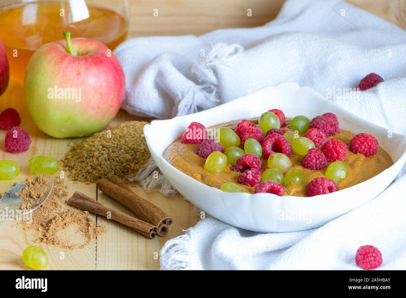Healthy meal concept background. Vegan flax porridge with raspberry and grape raisins in white bowl on fabric. Apple, honey, cinnamon sticks and Stock Photo