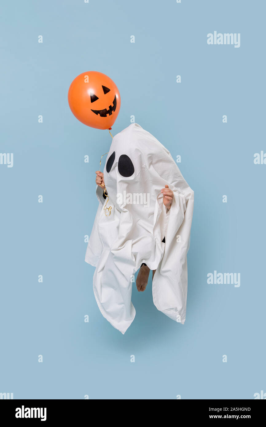 Little kid dressed as white ghost on halloween floating in the air over blue Stock Photo