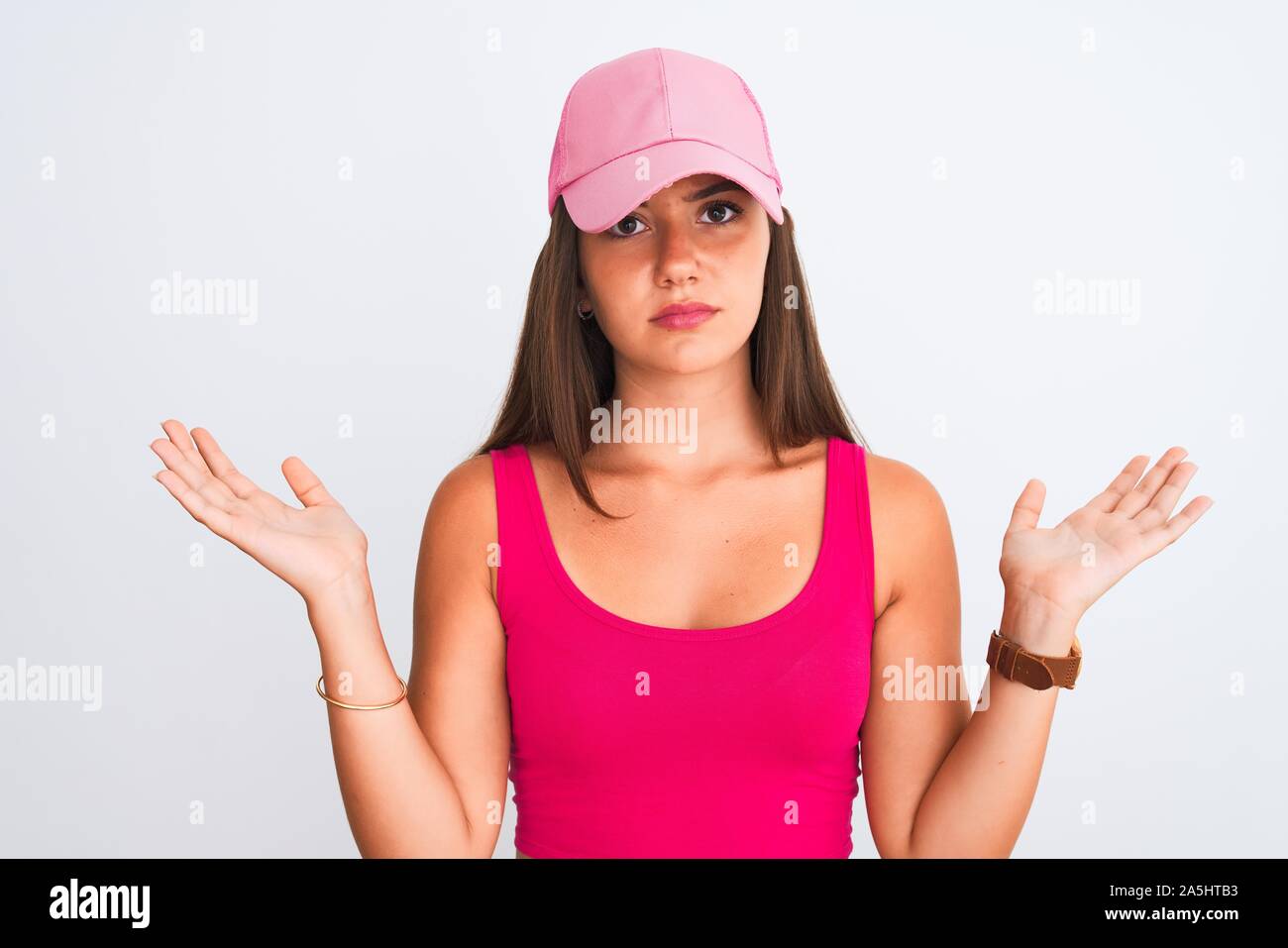 Young beautiful girl wearing pink casual t-shirt and cap over isolated white background clueless and confused expression with arms and hands raised. D Stock Photo