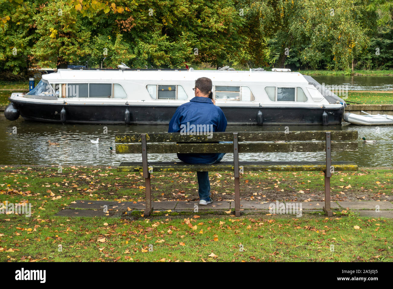 A man sits on a wooden bench next to The River Thames at Windsor while making a telephone call. Stock Photo