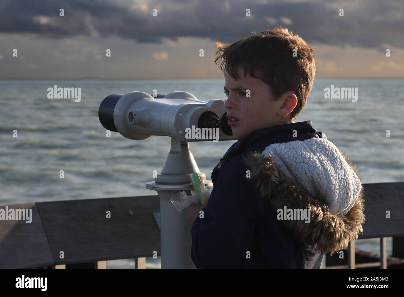 Young child looking to side next to large scale public binoculars on Hastings pier in daytime Stock Photo