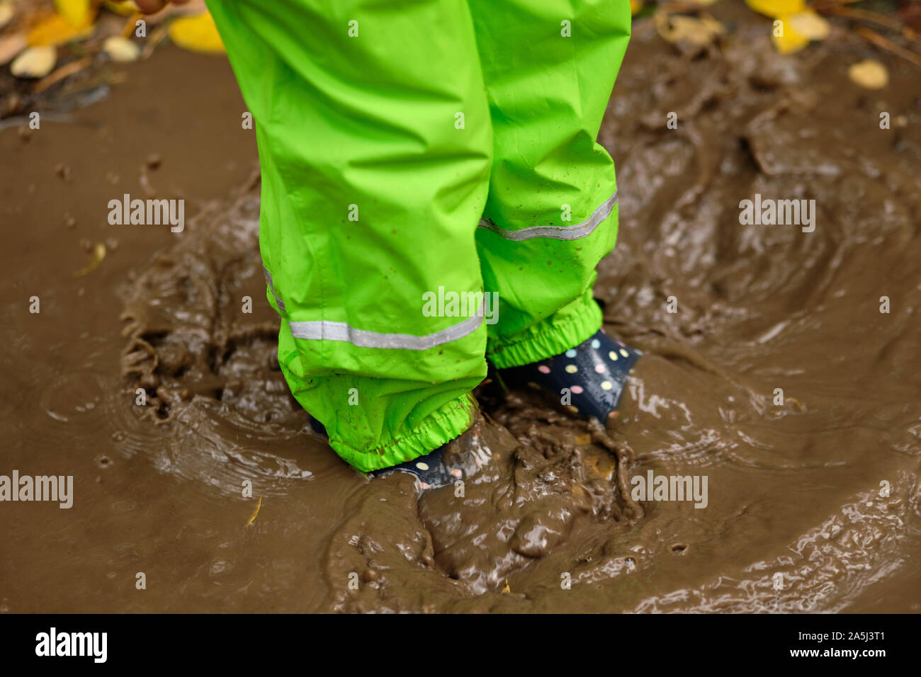 Low section of a child in green waterproof pants and rubber boots standing in a brown muddy puddle in the forest with fallen autumn leaves on a rainy Stock Photo