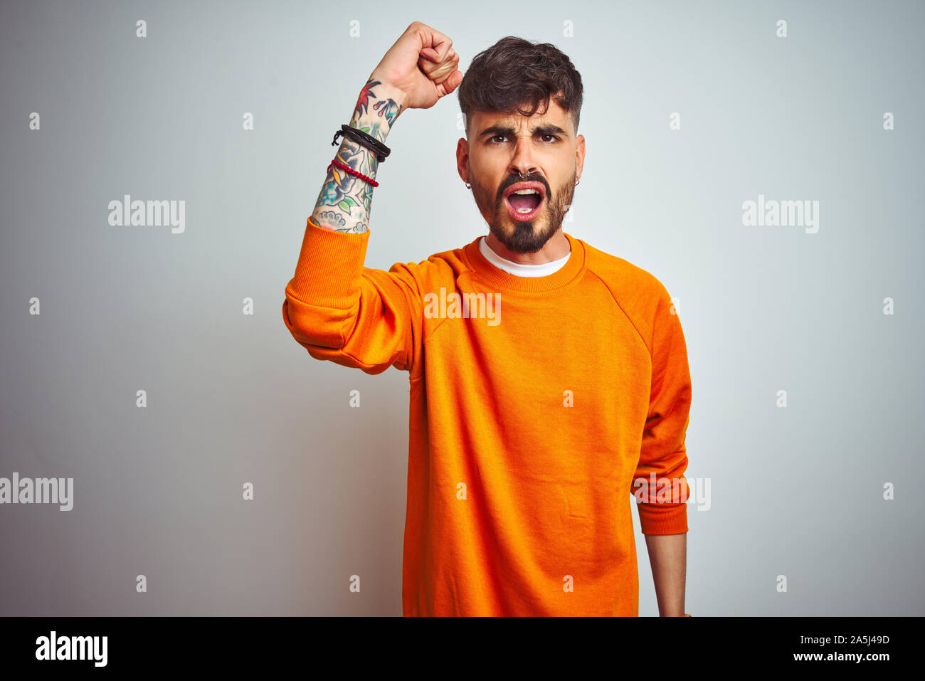 Young man with tattoo wearing orange sweater standing over isolated white background angry and mad raising fist frustrated and furious while shouting Stock Photo
