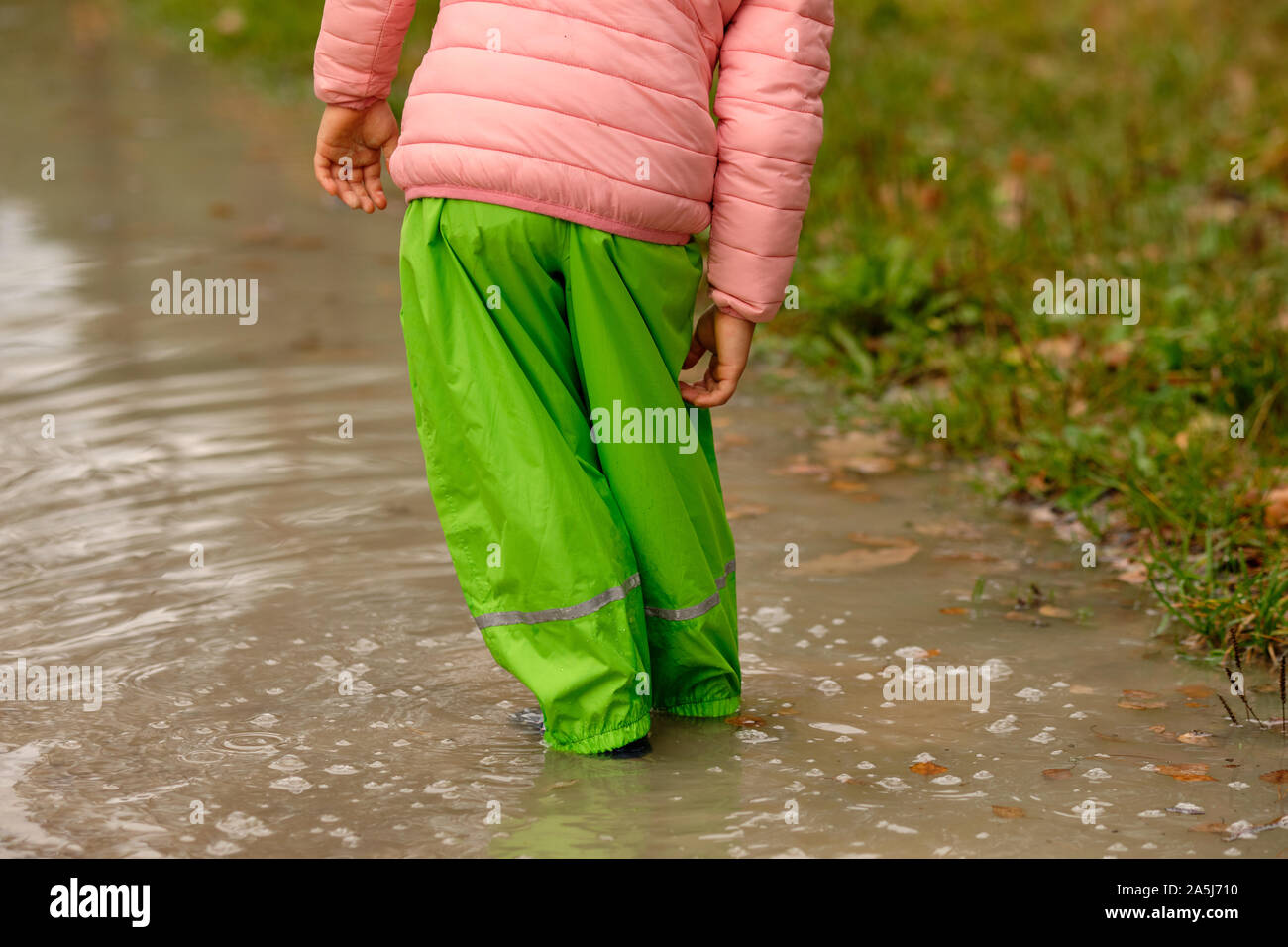 Low section of a child girl in green waterproof pants and rubber boots standing in a huge rain puddle in the forest on a rainy autumn day in October i Stock Photo
