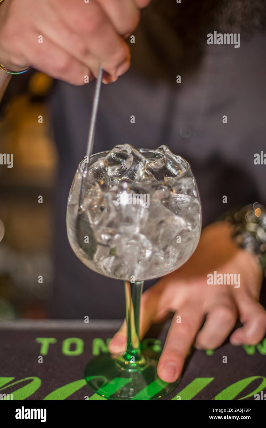 View of bartender preparing glass of tonic gin in flask with lots of ice in bar of drinks, blurred person as background Stock Photo