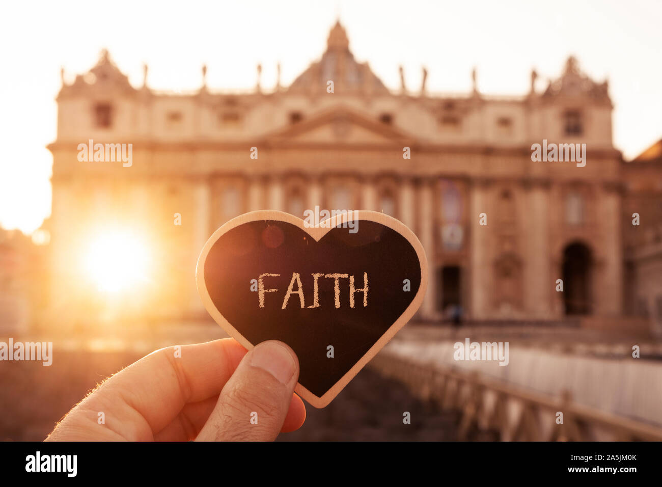 closeup of the hand of a caucasian man holding a black heart-shaped signboard with the word faith written in it in front of the Basilica of St Peter i Stock Photo