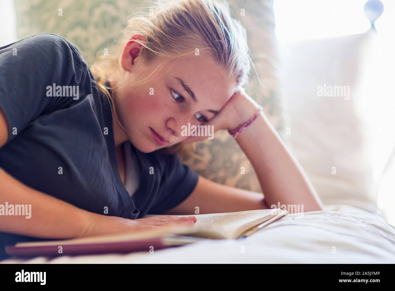 13 year old girl lying in her bed reading by window light Stock Photo