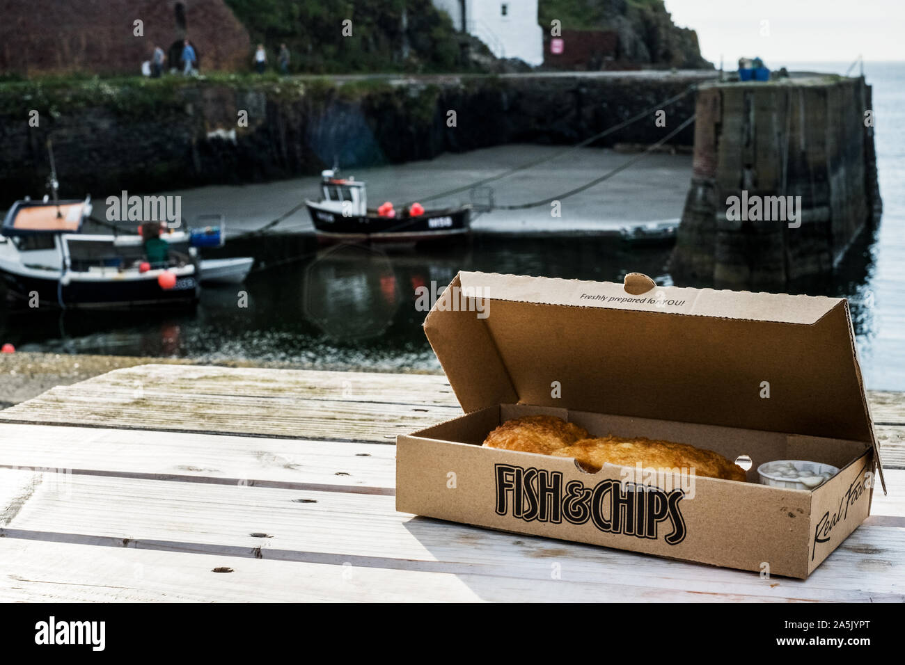 Box with fish and chips on a table in the small harbour of Porthgain on the Pembrokeshire Coast, Wales, UK. Stock Photo