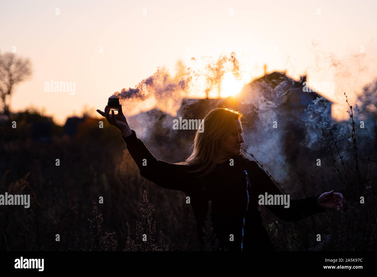 a woman with long hair holds a smoking bowl in her hand. portrait at sunset. allegory of magic, freedom, protest Stock Photo