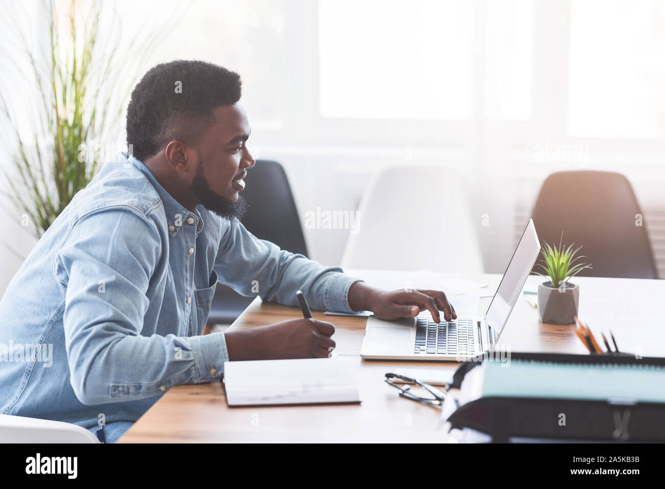 African worker taking notes while using laptop in office Stock Photo