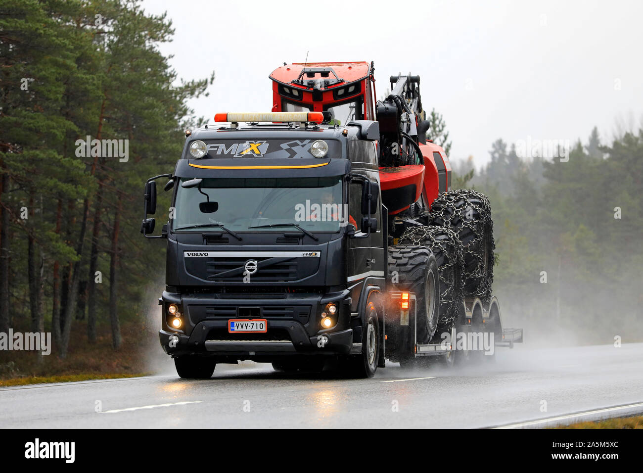 Black Volvo FMX of Metsakuljetus transports Komatsu 911 forest harvester on trailer on wet highway in autumn. Tenhola, Finland. October 18, 2019. Stock Photo