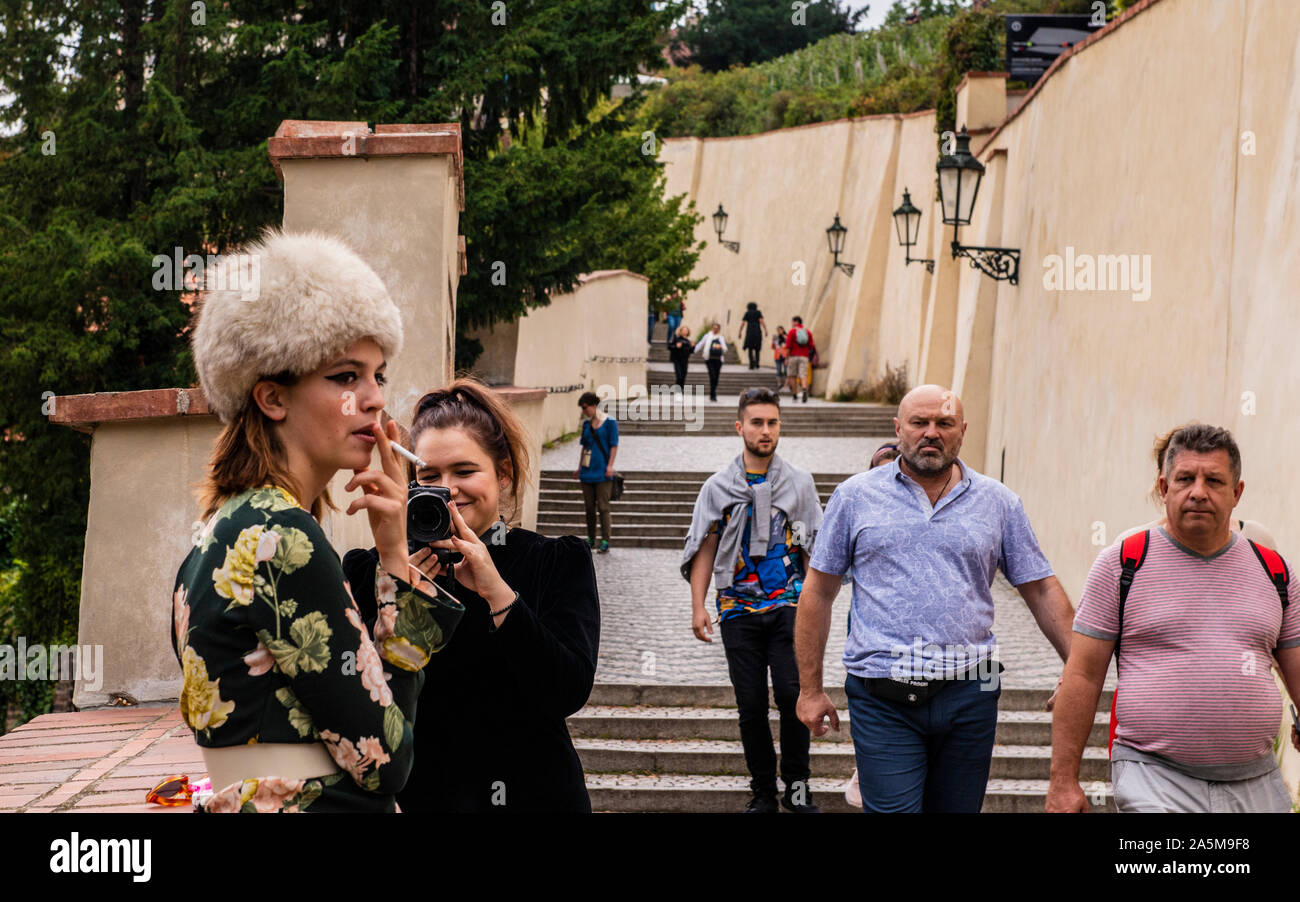 Woman taking photograph of fashionably dressed friend smoking on walkway, Prague, Czech Republic Stock Photo