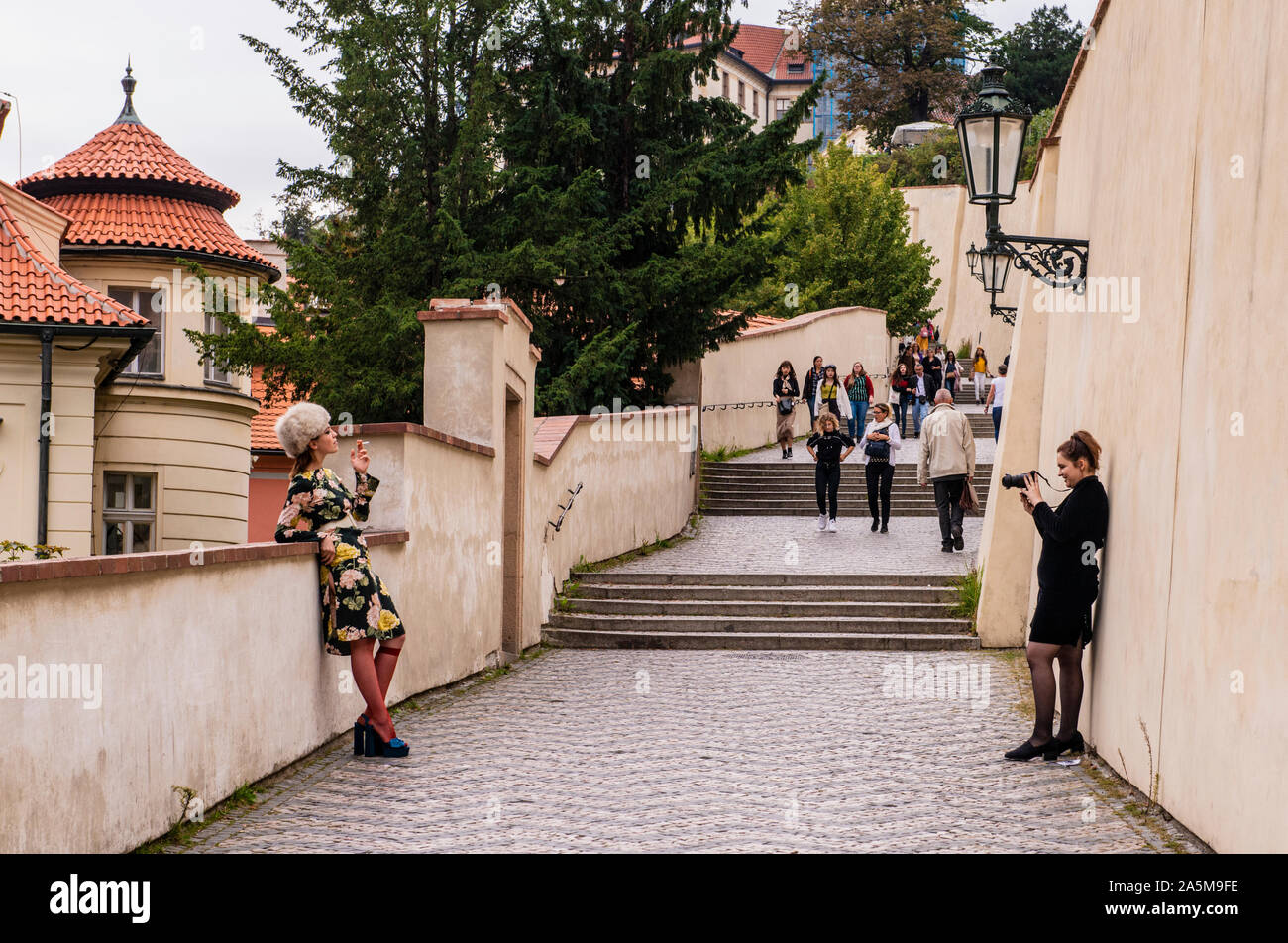 Woman taking photograph of fashionably dressed friend smoking on walkway, Prague, Czech Republic Stock Photo