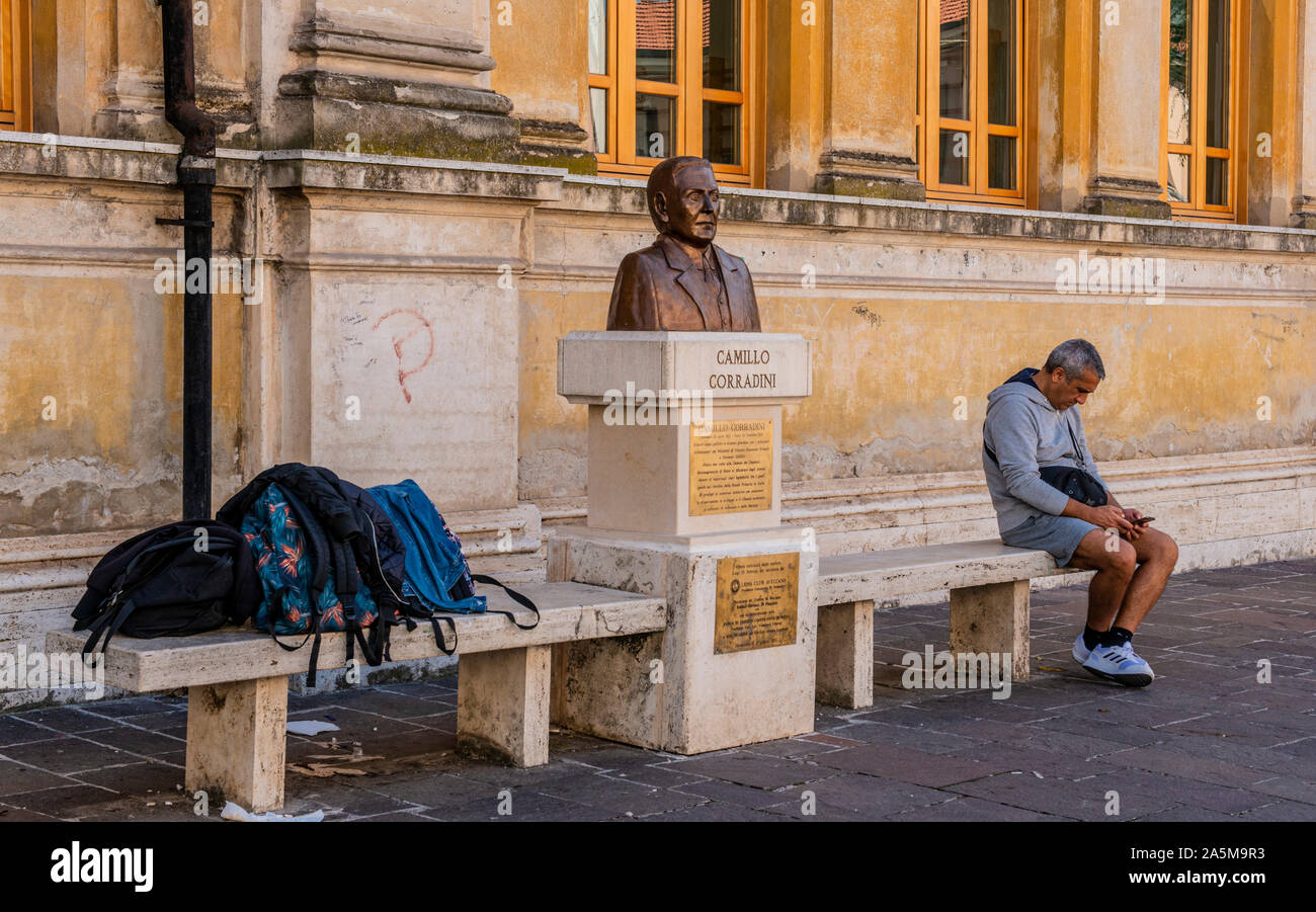 Man using smartphone beside bust sculpture of Camillo Corradini, Piazza Risorgimento, Avezzano, Abruzzo, Italy Stock Photo