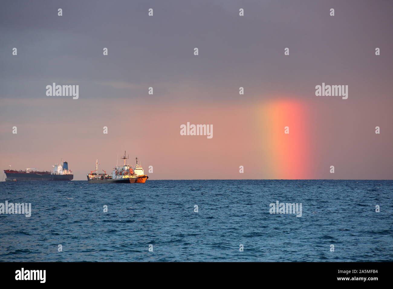 beautiful unusual short rainbow on sea horizon, next to two ships / boats, cover by heavy grey rainy clouds Stock Photo