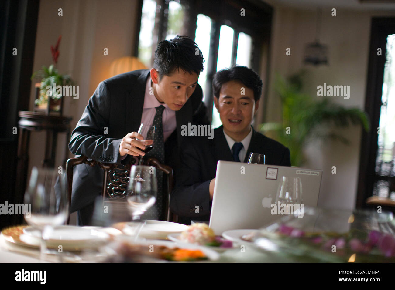 Two businessmen using a laptop while drinking wine at a table inside a restaurant. Stock Photo