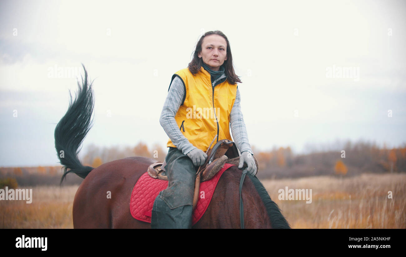 A woman in yellow vest is sitting on a bay horse - outdoors Stock Photo