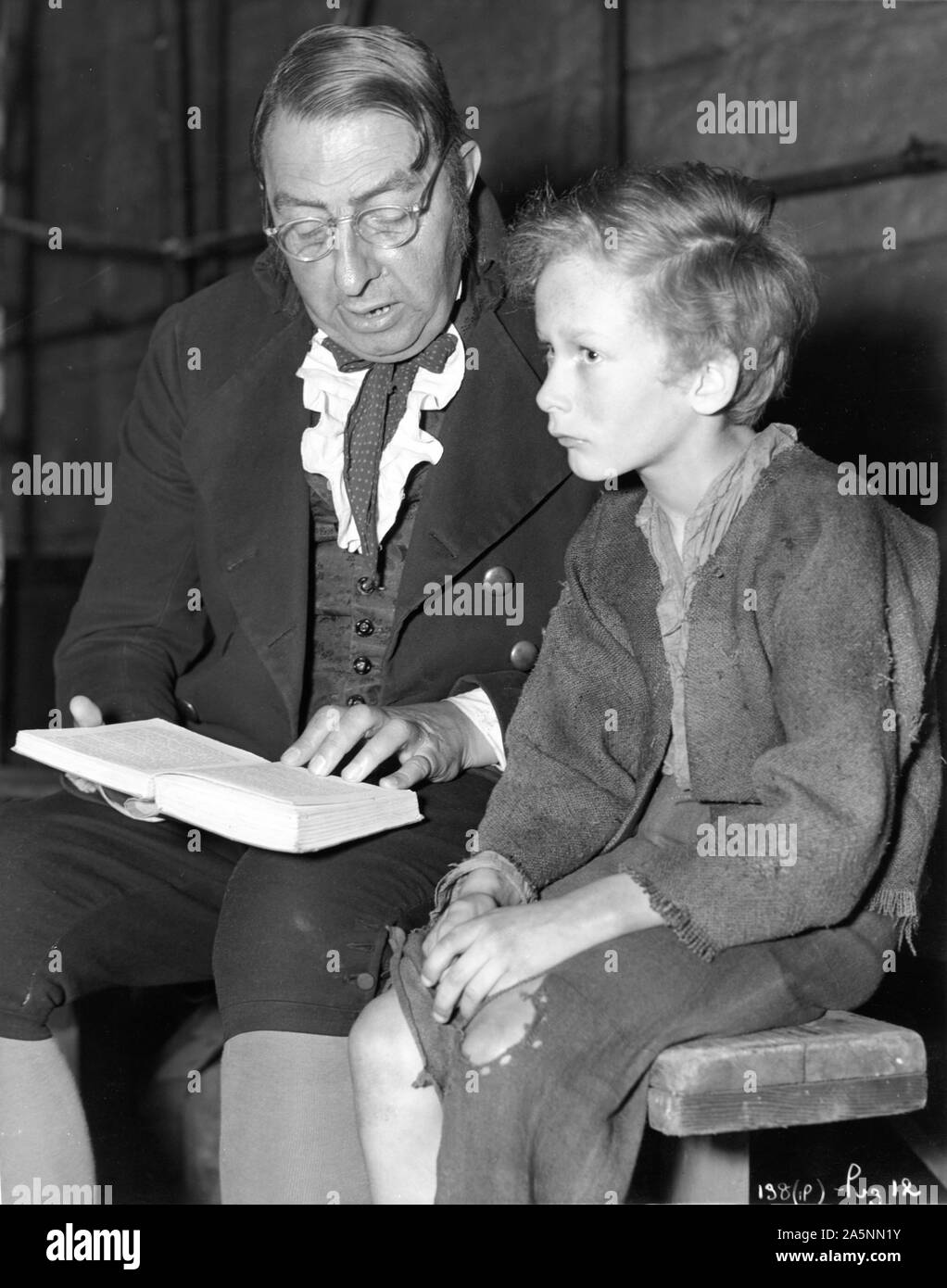 KENNETH DOWNY as the Workhouse Master and JOHN HOWARD DAVIES as OLIVER TWIST 1948 director DAVID LEAN novel CHARLES DICKENS on set candid during filming reading the original book Photo by Charles Trigg Cineguild / General Film Distributors (GFD) Stock Photo