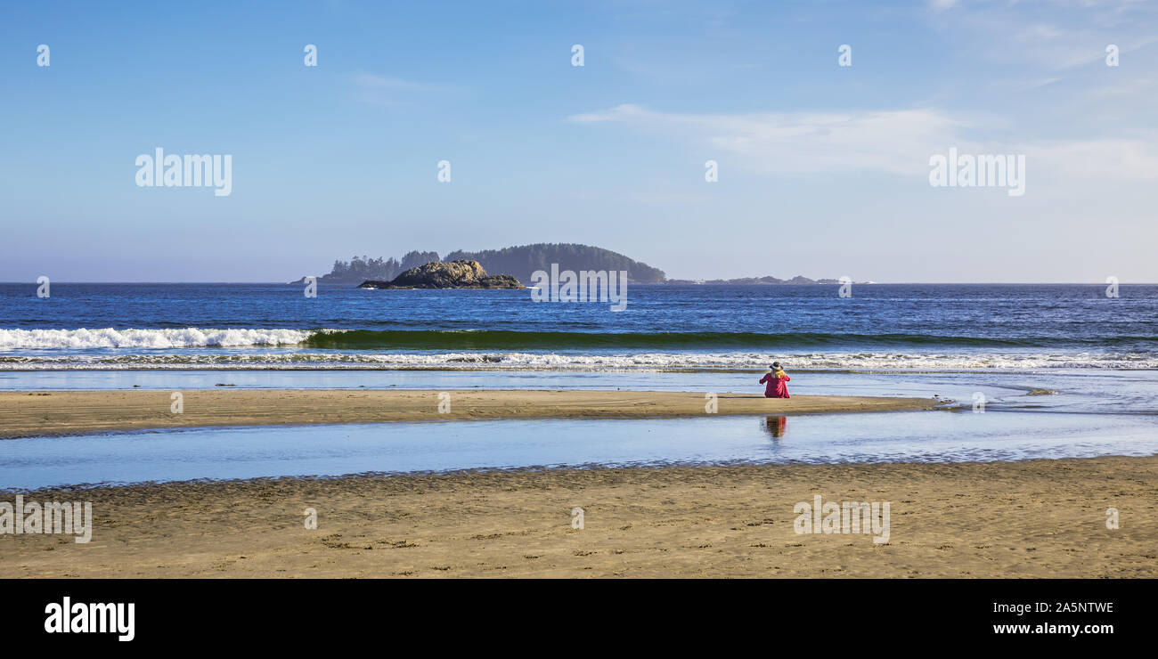 View of woman with hat and red shirt sitting alone at the beach staring at the west pacific ocean near Tofino on Vancouver Island Stock Photo