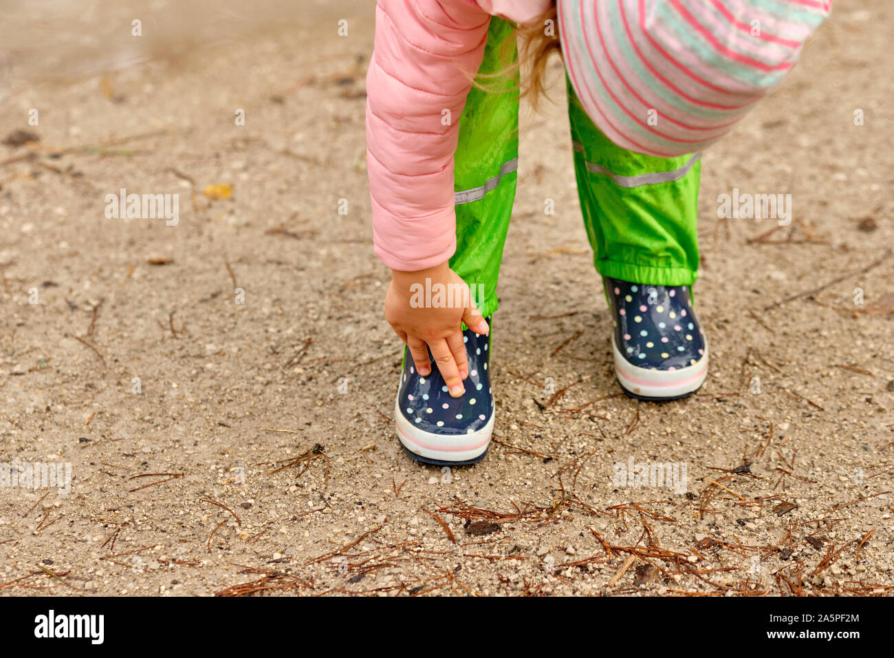 A child in green waterproof pants and spotted rubber boots standing on a wet gravel road in the forest and touching her boots with her fingers on a ra Stock Photo