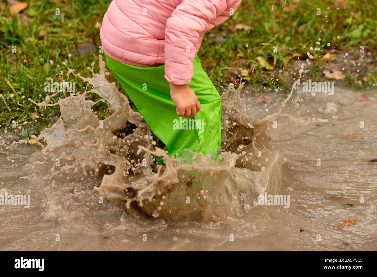 Low section of a child girl in green waterproof pants and rubber boots jumping into a massive rain puddle with the water splashing high up in the air Stock Photo