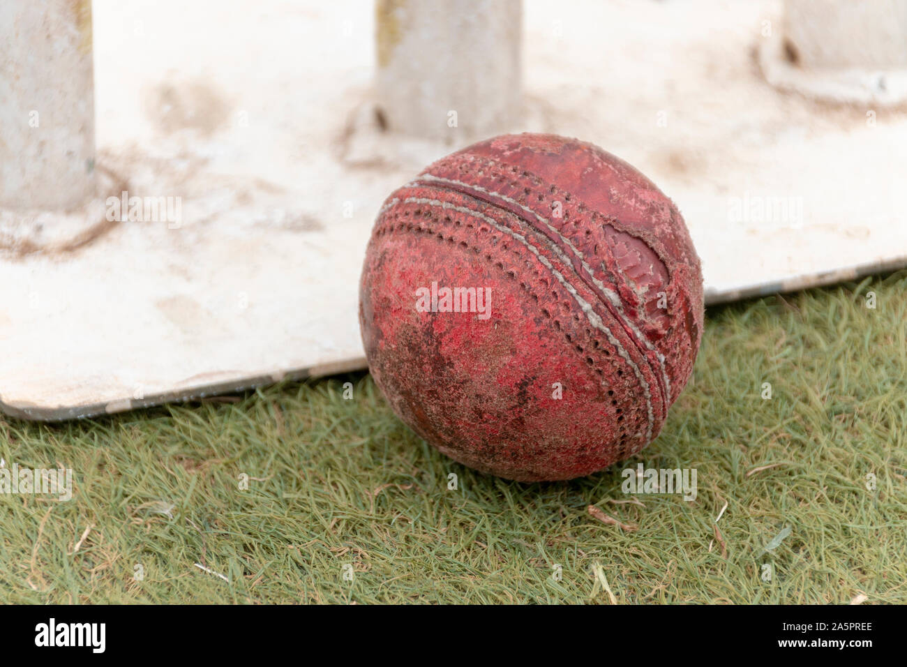 A close up view of a old well used red cricket ball on a grass pitch next to white metal wickets Stock Photo