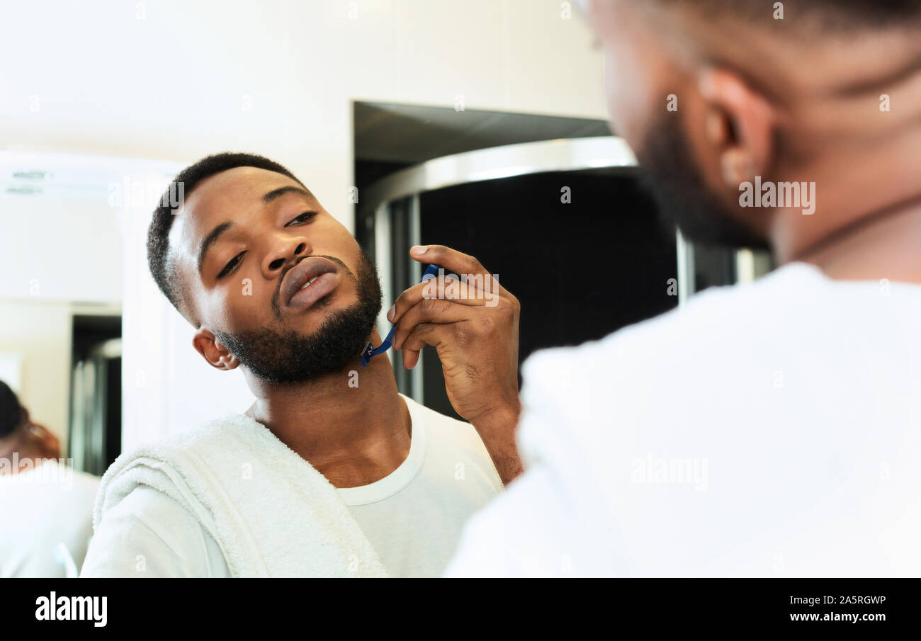 Young black man shaving beard, looking at bathroom mirror Stock Photo