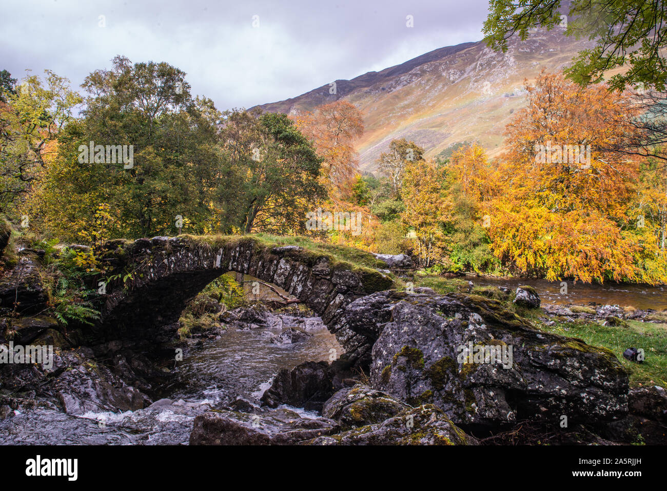 Old curved Roman Bridge in Glen Lyon, Scottish Highlands with rainbow on hills in background Stock Photo