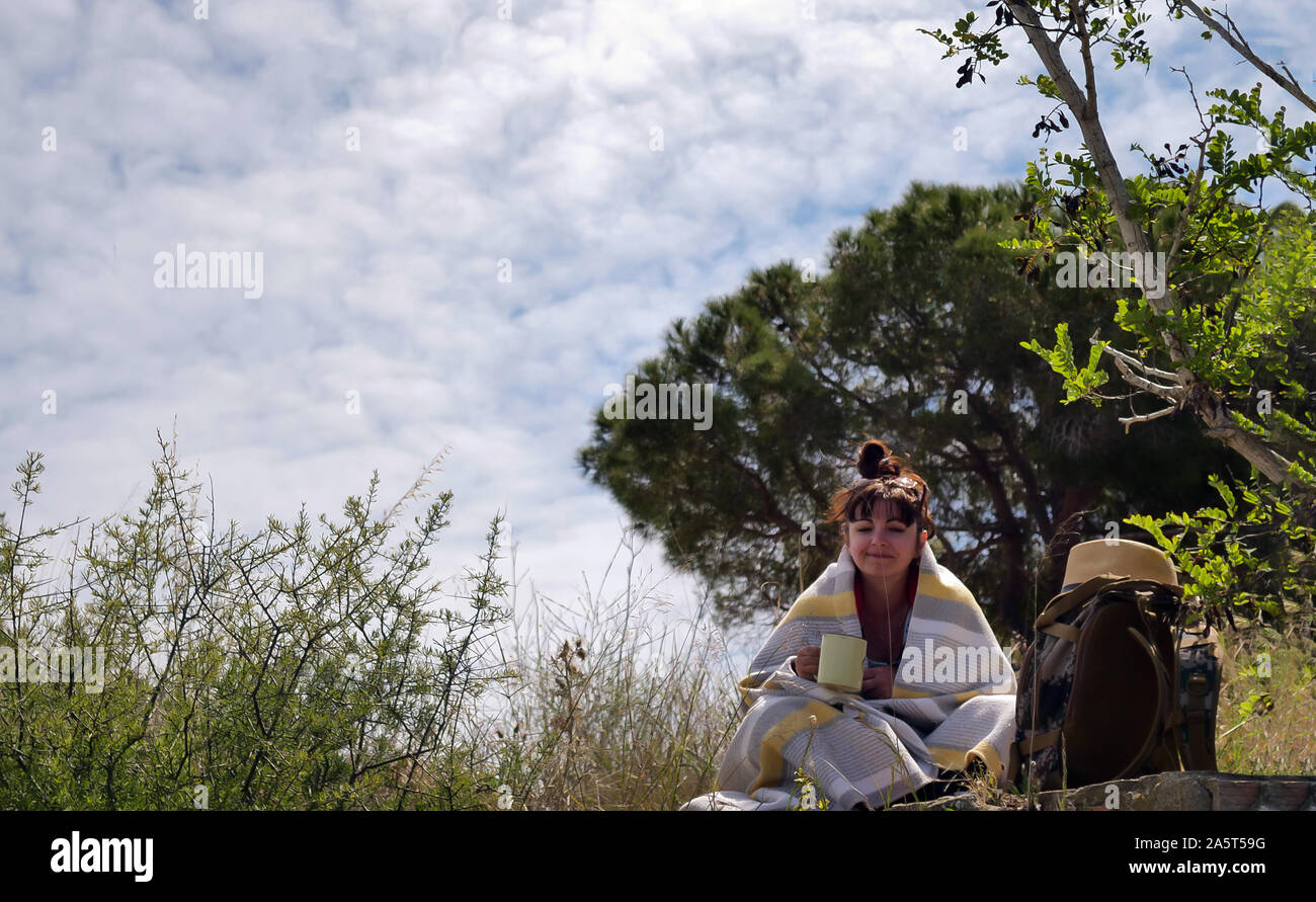 Nice girl tourist sitting in nature wrapped in a blanket with a mug of hot drink Stock Photo