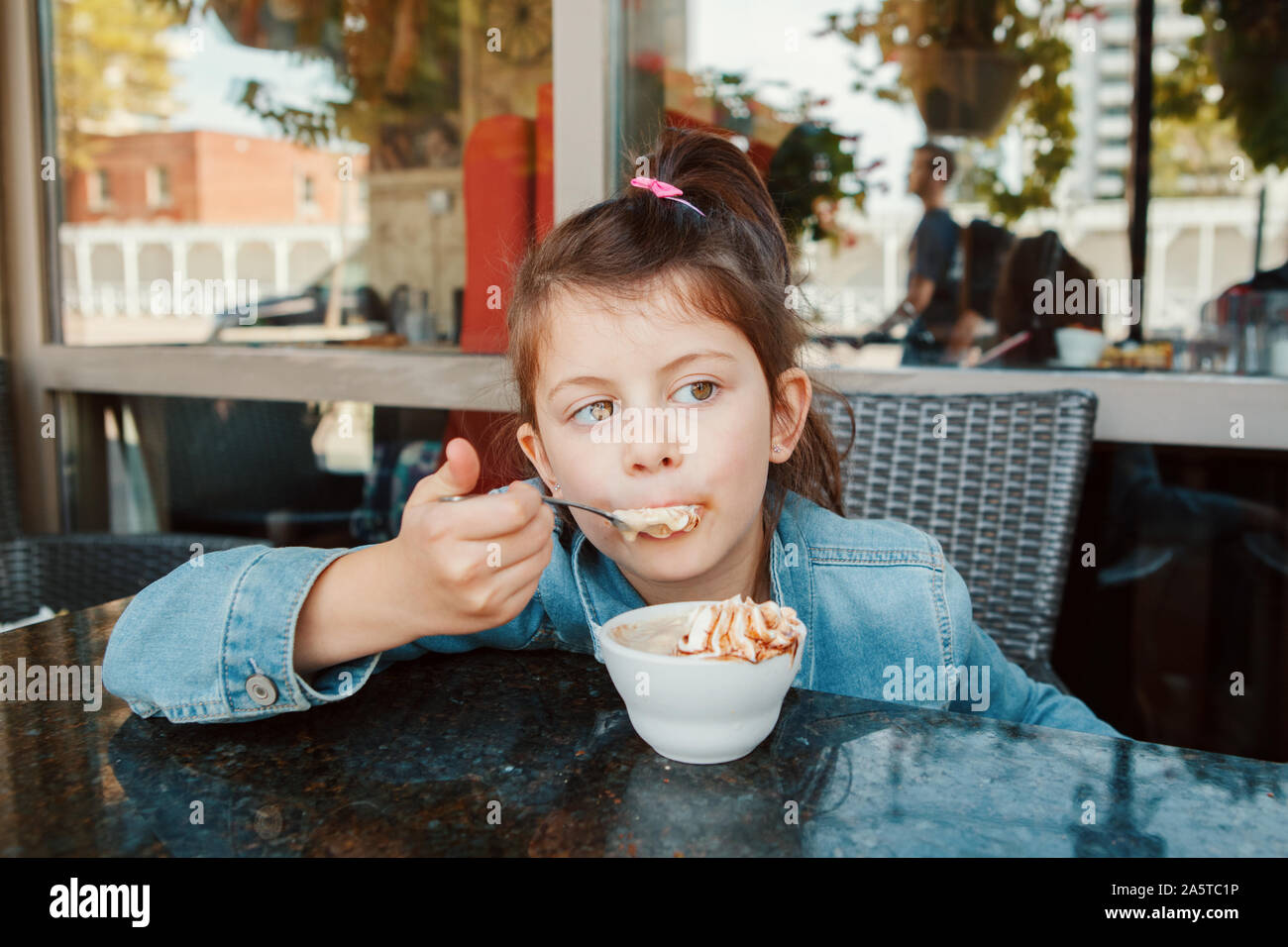 Funny Caucasian preschool girl eating sweet dessert with spoon in cafe. Child kid having fun in restaurant patio enjoying food drink. Happy authentic Stock Photo