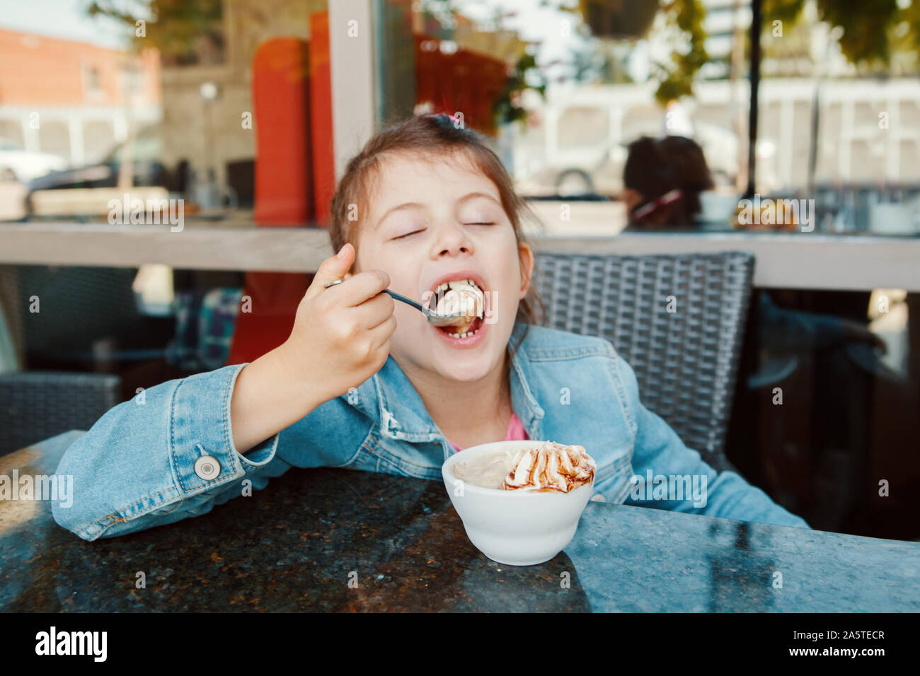 Funny Caucasian preschool girl eating sweet dessert with spoon in cafe. Child kid having fun in restaurant patio enjoying food drink. Happy authentic Stock Photo