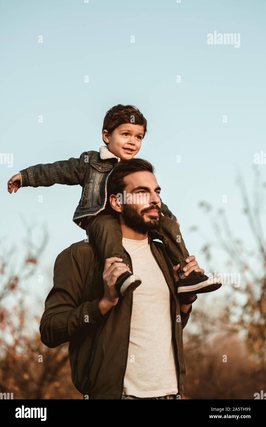 Little boy sitting on father's shoulders on blue sky background. Stock Photo