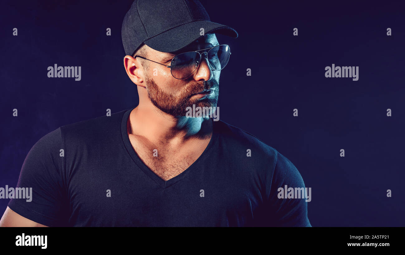 Portrait of brutal bearded man in sunglassesand baseball cap at dark photo studio. Stock Photo