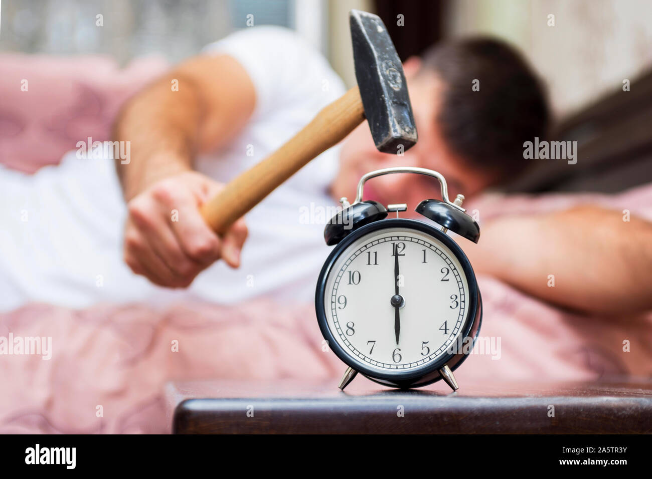 Time to wake up. Lazy man tries to break the alarm clock with hammer from the bed. Stock Photo