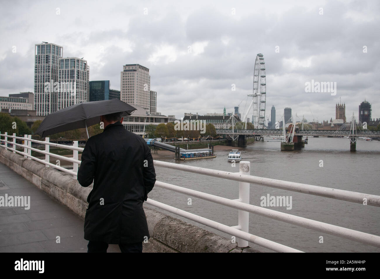Man in a black coat holding a black umbrella against the wind and rain walks arcoss a bridge over the river thames with the South Bank and London Eye. Stock Photo