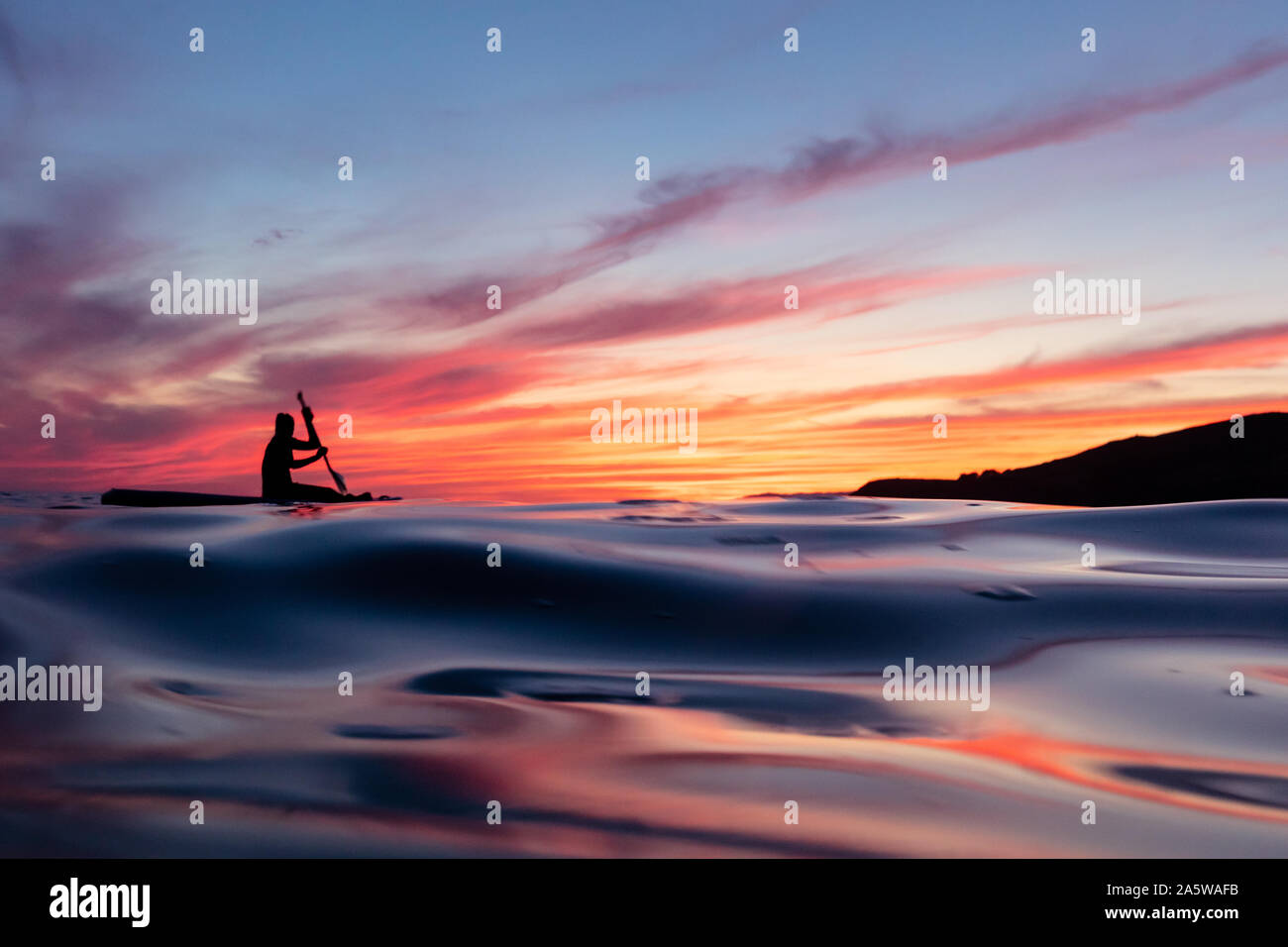 A woman paddles across the ocean surface reflecting vibrant sunset afterglow from colorful clouds. Stock Photo