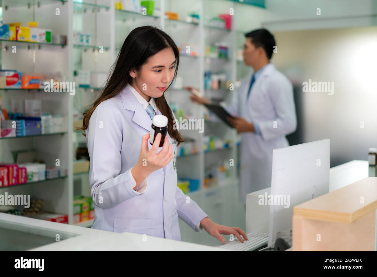 Confident Asian young female pharmacist with smile holding a medicine bottle and searching that product in the computer database in the pharmacy drugs Stock Photo