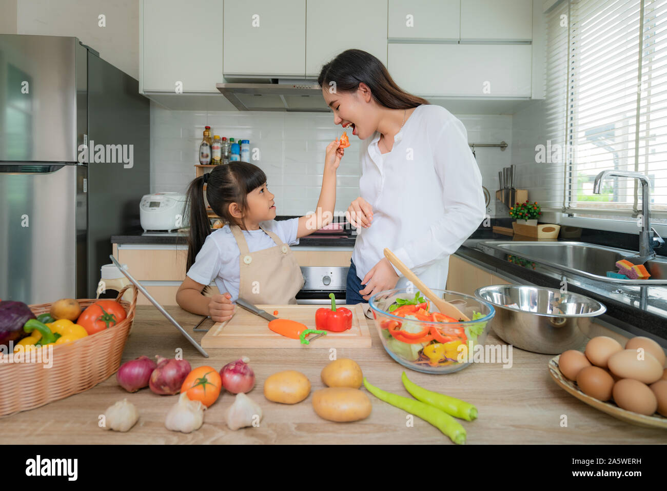 Portrait of beautiful Asian young woman and her daughter cooking salad for lunch and feeding tomato to her mother while making food, Family life love Stock Photo