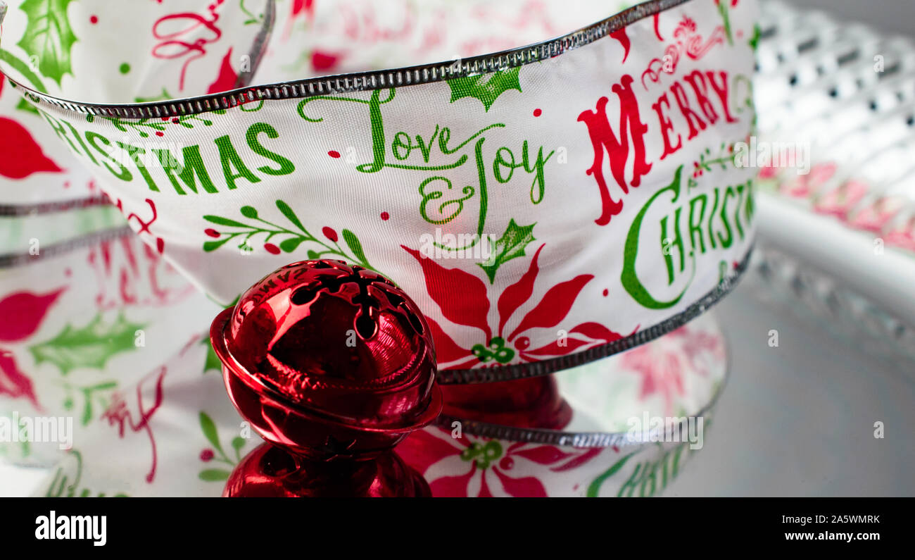 Red Jingle Bell and Decorative Merry Christmas Ribbon with the Words Love & Joy in Red and Green on Silver Reflective Surface Stock Photo