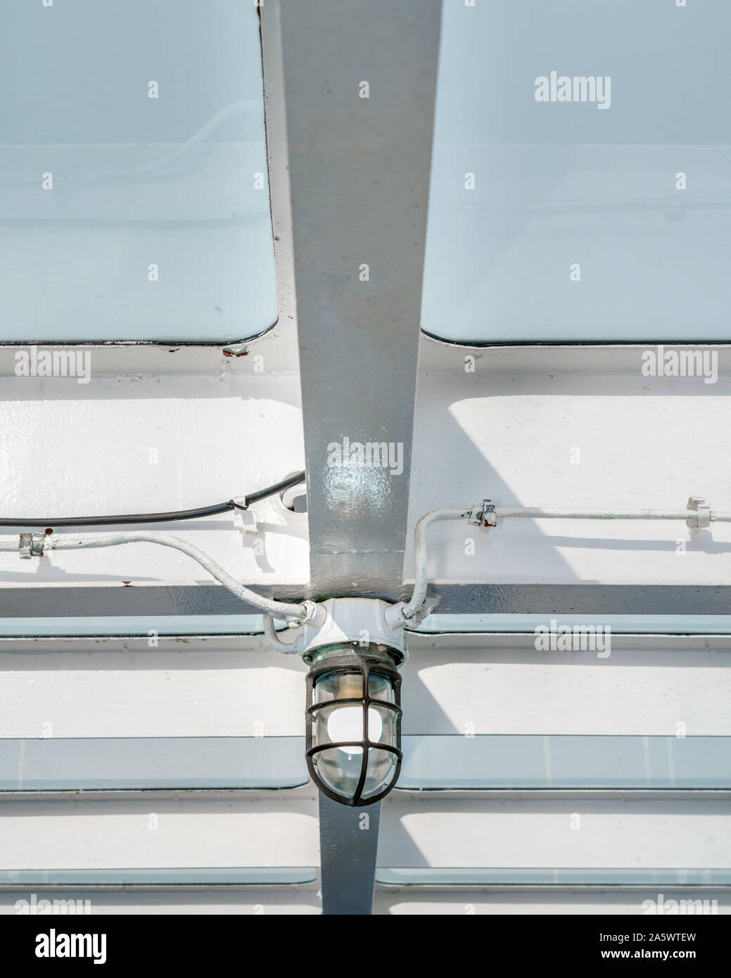 Detail of marine light fixture from underneath, attached to painted white metal beams aboard a ship with view of pale blue sky through ceiling panels. Stock Photo