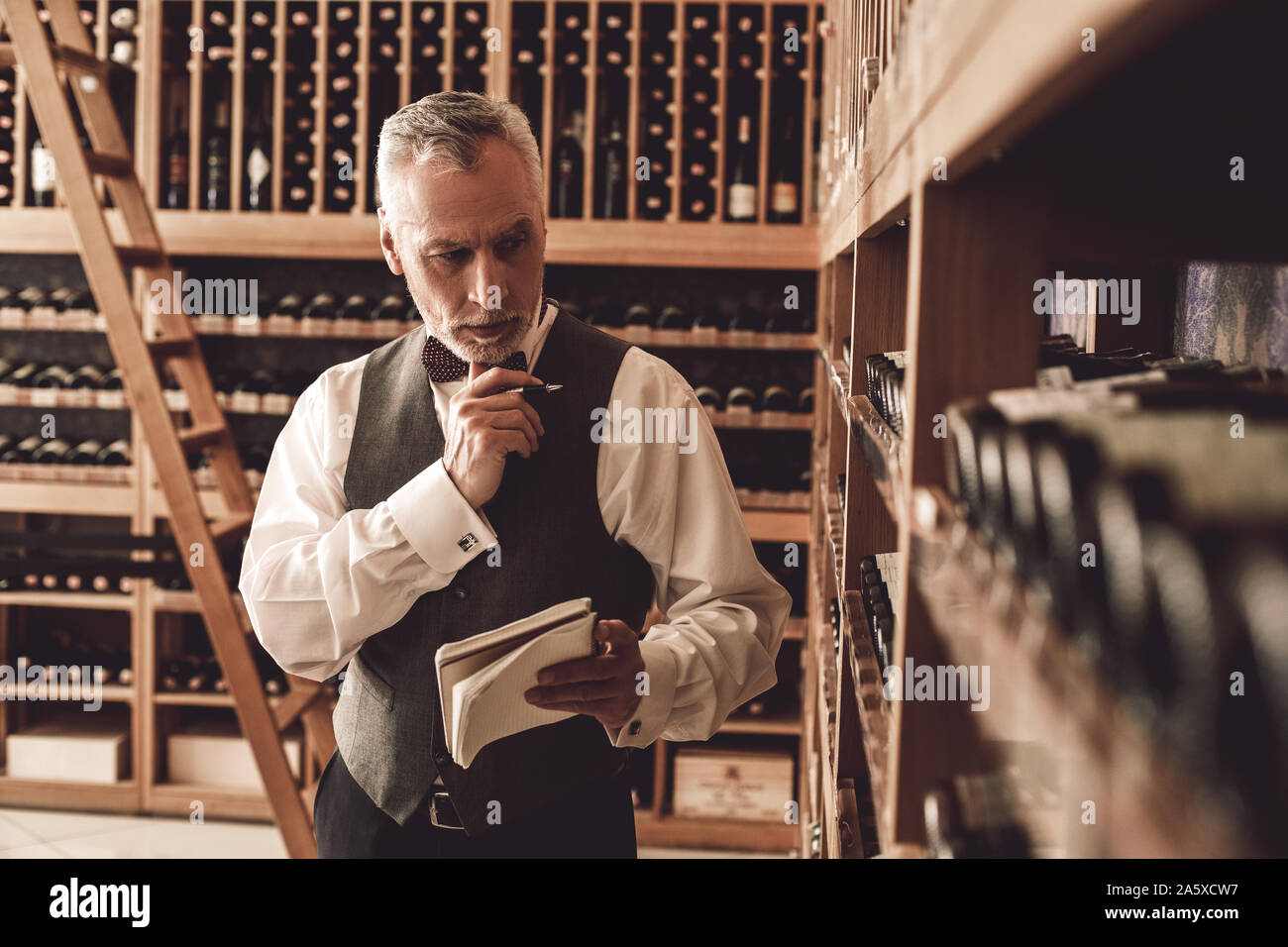 Sommelier Concept. Senior man standing with notebook checking wine collection pensive Stock Photo