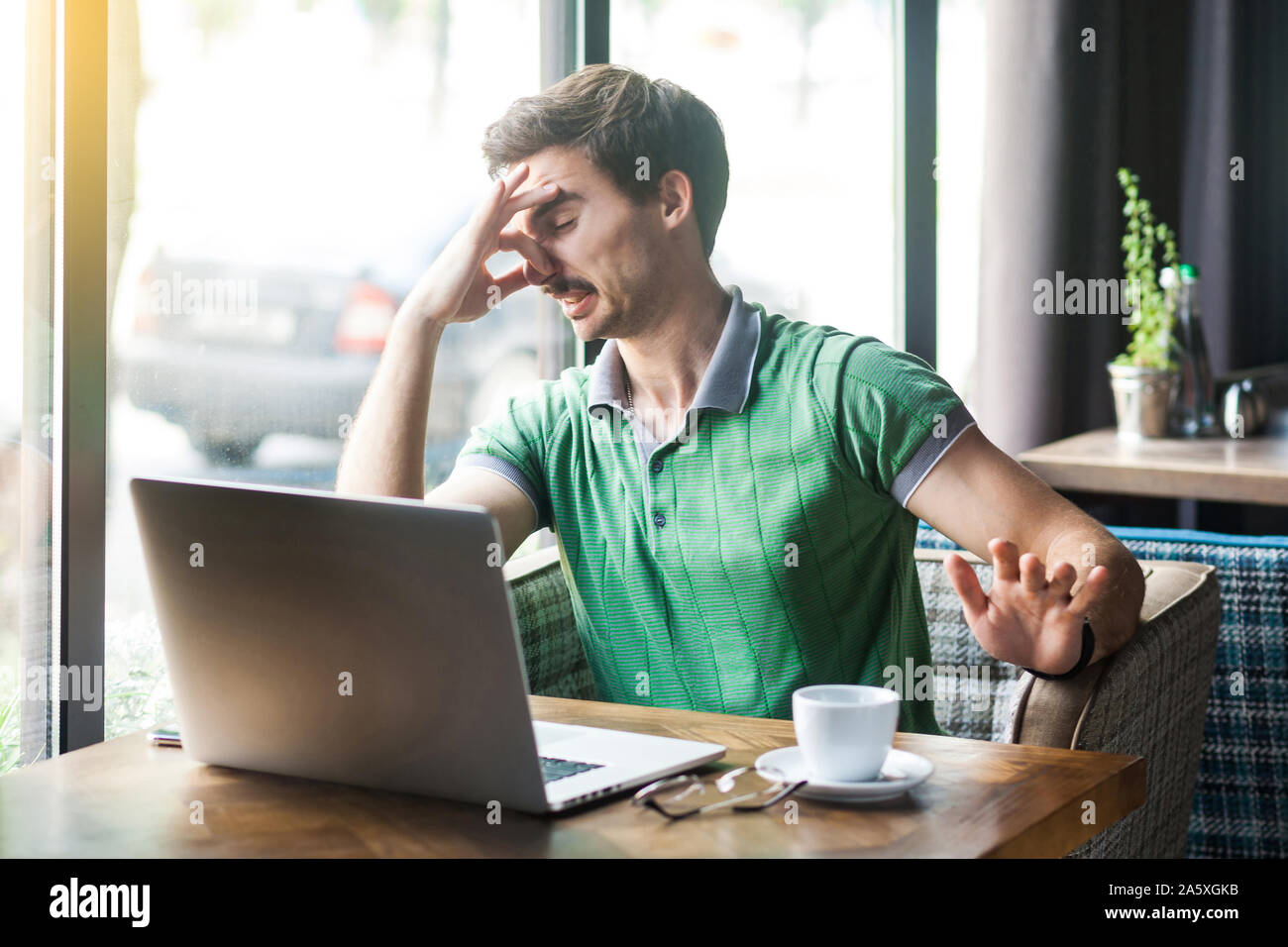 Bad smell. Young dissatisfied businessman in green t-shirt sitting and working on laptop, pinching his nose with negative emotion. business and freela Stock Photo