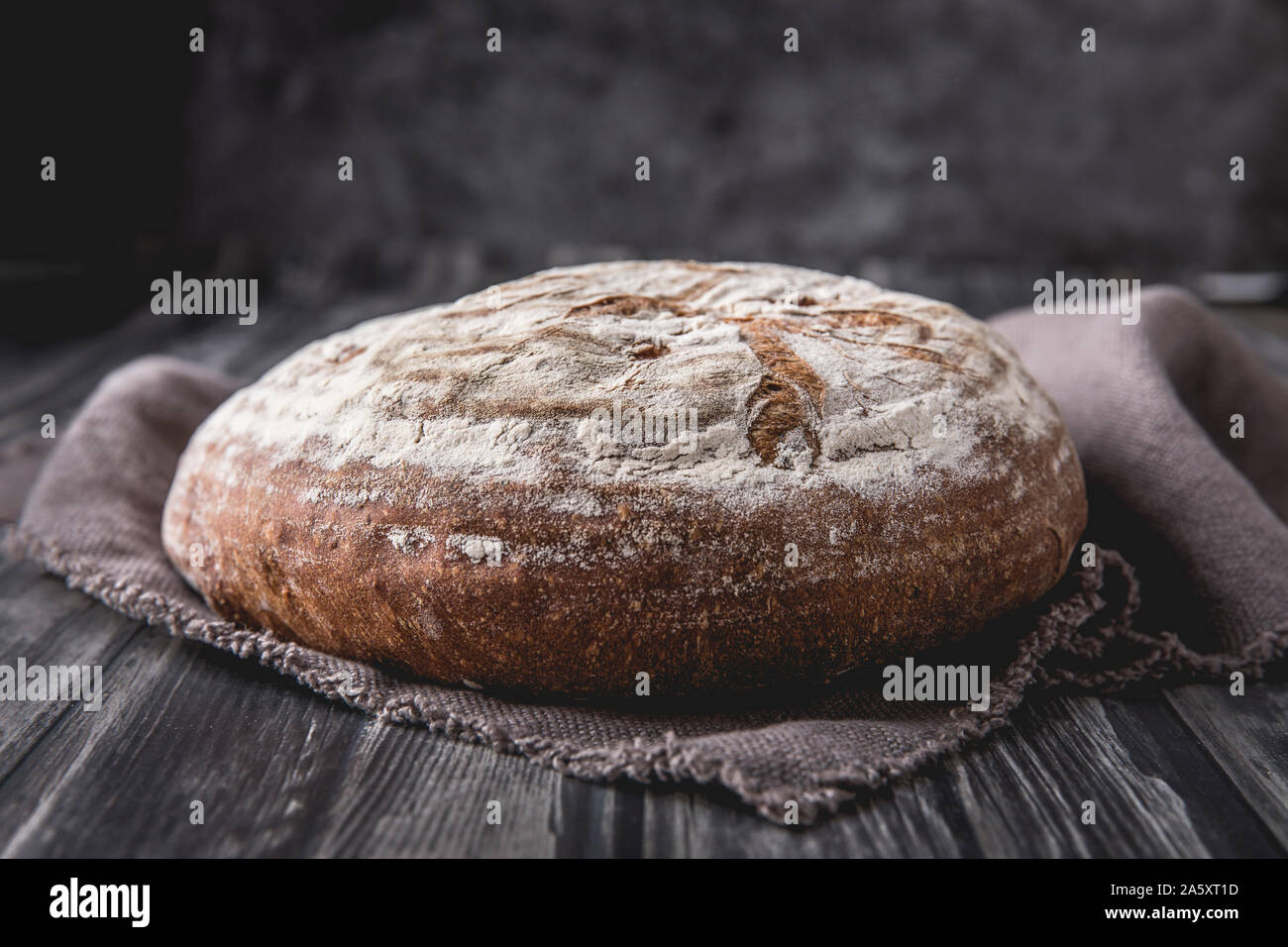 A homemade round levain bread with rye and wheat flour. that is laid on a dark wooden table, with a dark background. There is a brown soft linen cloth Stock Photo