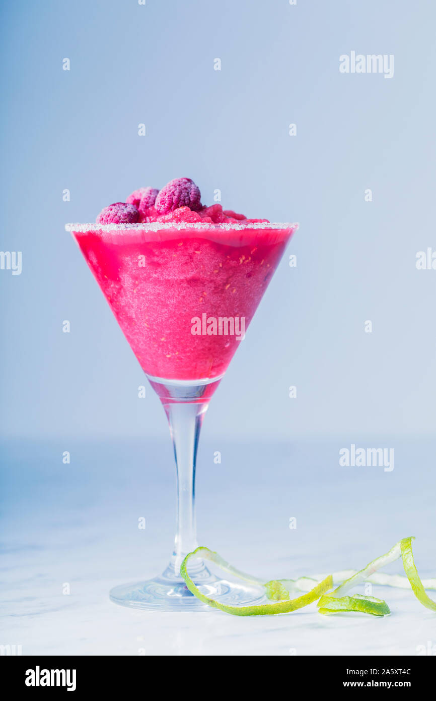 A fresh pink frozen raspberry drink in a sugar rimmed martini glass. The drink is decorated with frozen raspberries and a lime zest peel. The backgrou Stock Photo