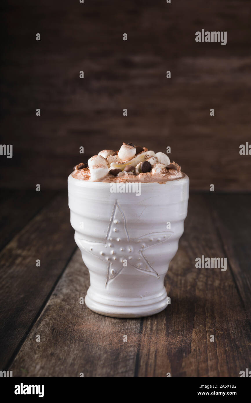 A white mug with luxurious hot chocolate with whipped cream and pieces of marshmallows and chocolate chips. Horizontal shot slightly from above. Minim Stock Photo