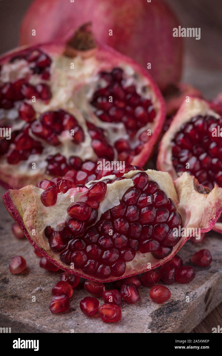 Pomegranate halved, slice and whole. The fruits are on a rustic stone surface and there are also some pomegranate seeds on the stone surface. Vertical Stock Photo