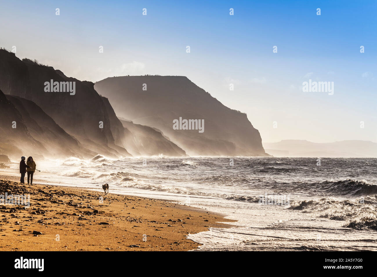 Two women walking their dog on Charmouth Beach at sunrise. Stock Photo